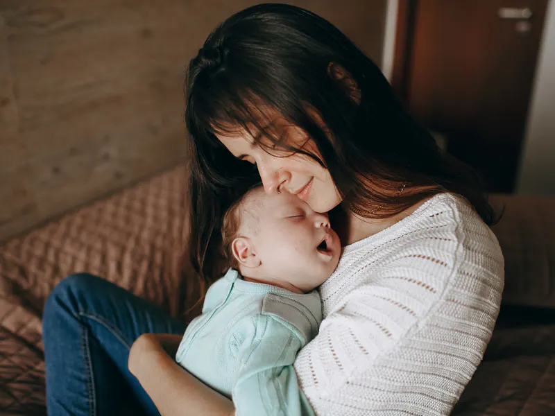 Imagem ilustrativa: mãe de olhos fechados beijando a cabeça do bebê recém-nascido que dorme no colo dela